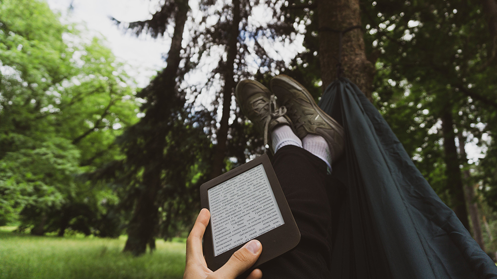 Man lying in a hammock in nature and reading a book on Kindle